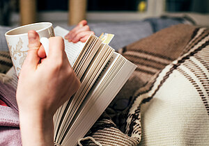 Kvinna sitter i soffan med en bok och en kopp te.